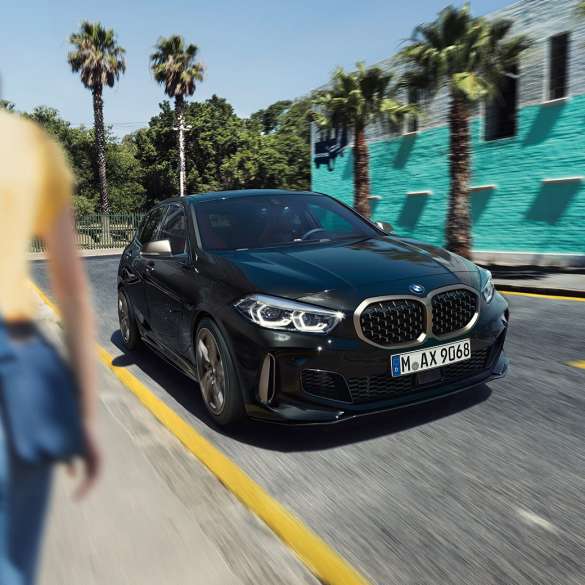 BMW M135i xDrive, מבט 3/4 קדמי. ה-BMW M135i xDrive בצבע שחור מטאלי Sapphire, מבט 3/4 בנהיגה, עם אישה ברקע.