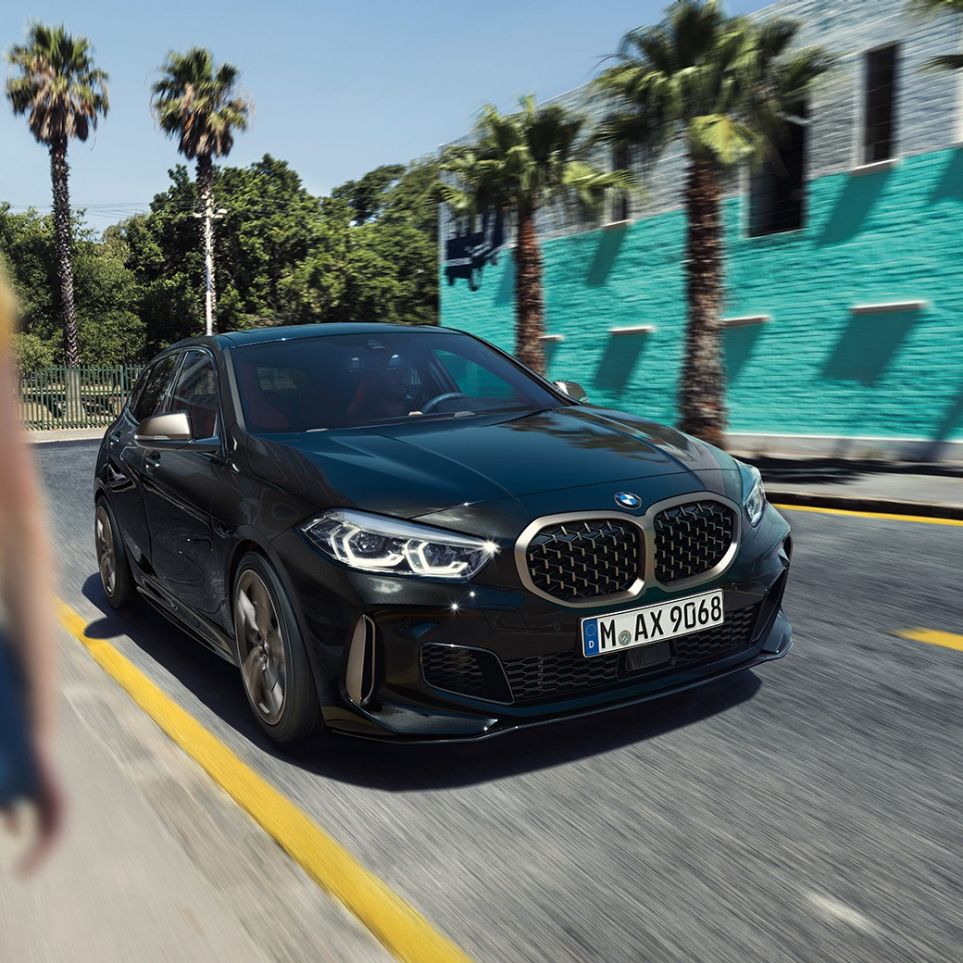 BMW M135i xDrive, מבט 3/4 קדמי. ה-BMW M135i xDrive בצבע שחור מטאלי Sapphire, מבט 3/4 בנהיגה, עם אישה ברקע.