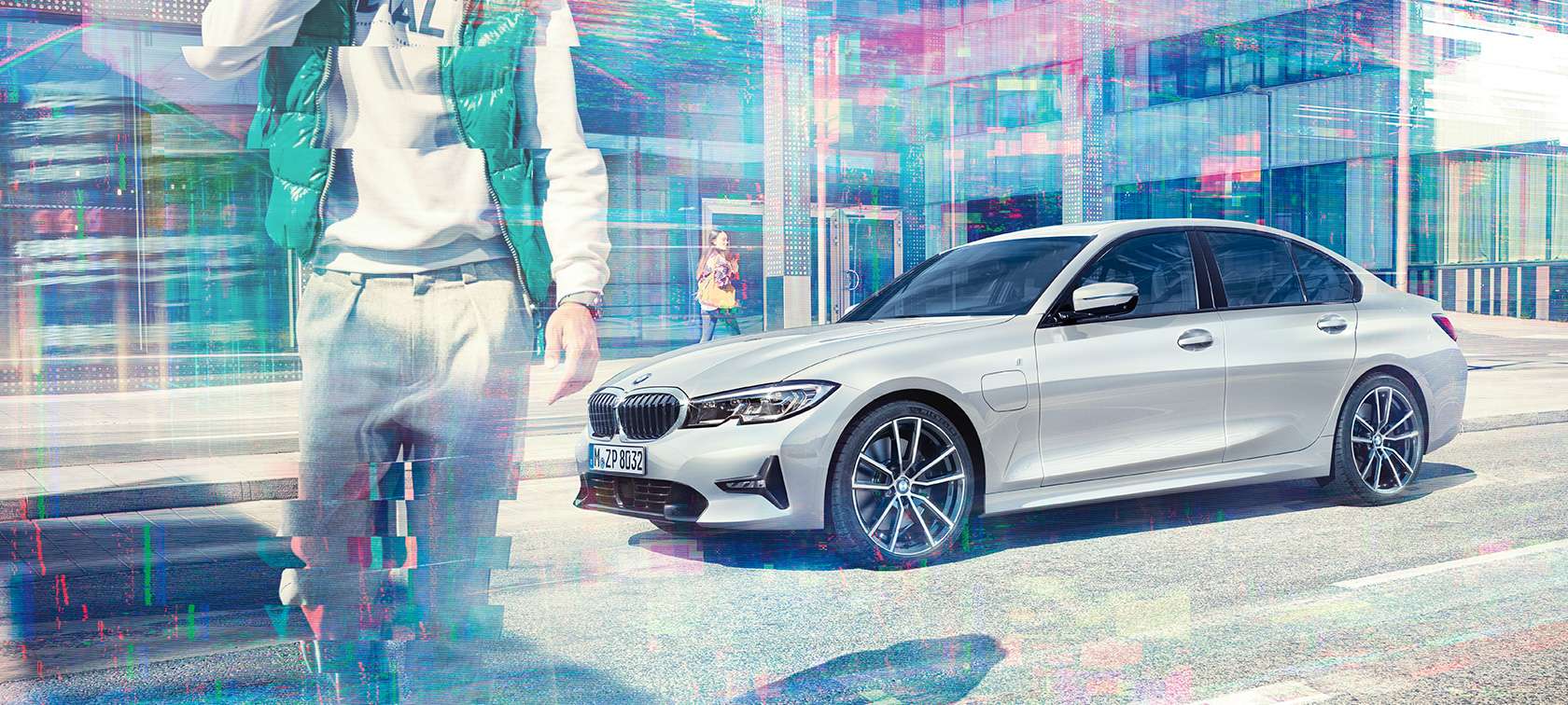 BMW סידרה 3, תצלום במבט צד של BMW 330e. תצלום במבט אלכסון-צד של BMW 330e על האספלט, על רקע של מבנה זכוכית בסביבה עירונית עם הולכי רגל.