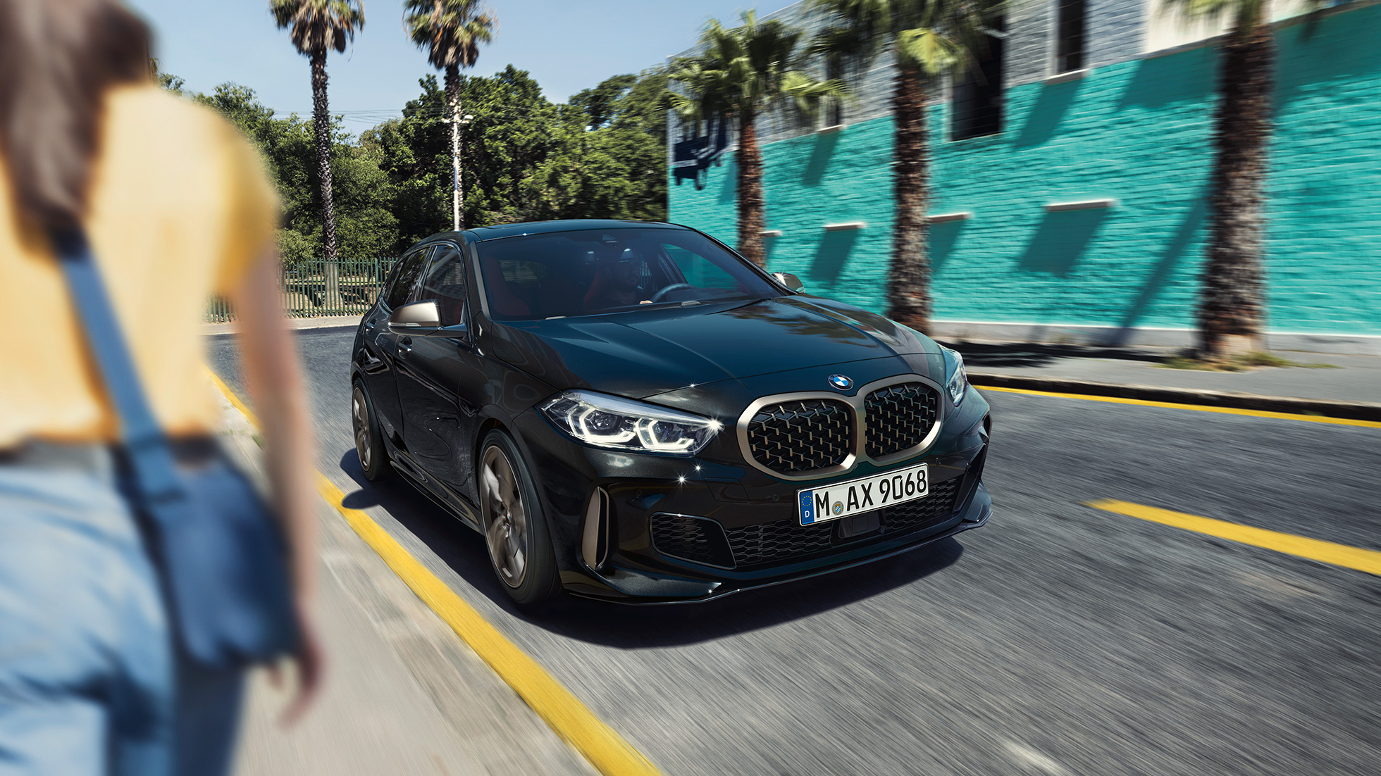 BMW M135i xDrive, מבט 3/4 קדמי. ה-BMW M135i xDrive בצבע שחור מטאלי Sapphire, מבט 3/4 בנהיגה, עם אישה ברקע.