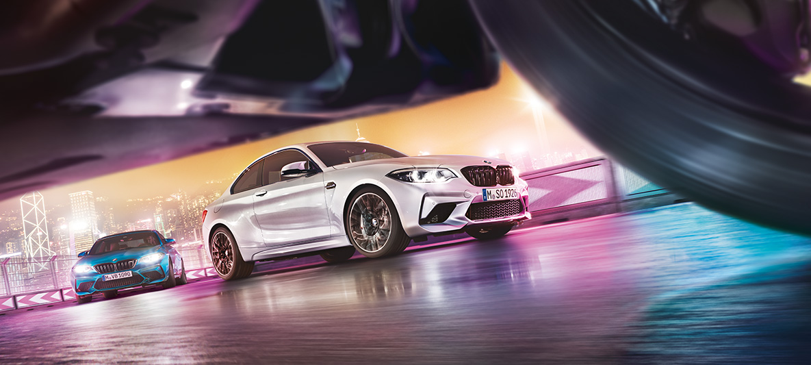 BMW M2 Competition Two BMW M2 Competition on race track in front of nocturnal cityscape in street-level view.